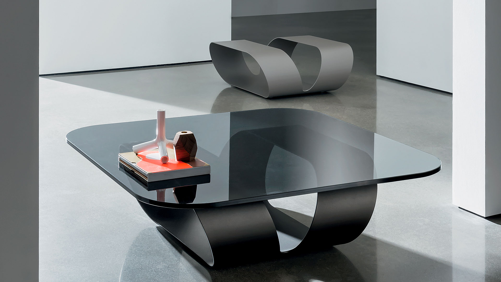 Tavolino in vetro sagomato quadrato con piano in vetro fumè e struttura composta da un'unica lastra di metallo curvata di colore grigio