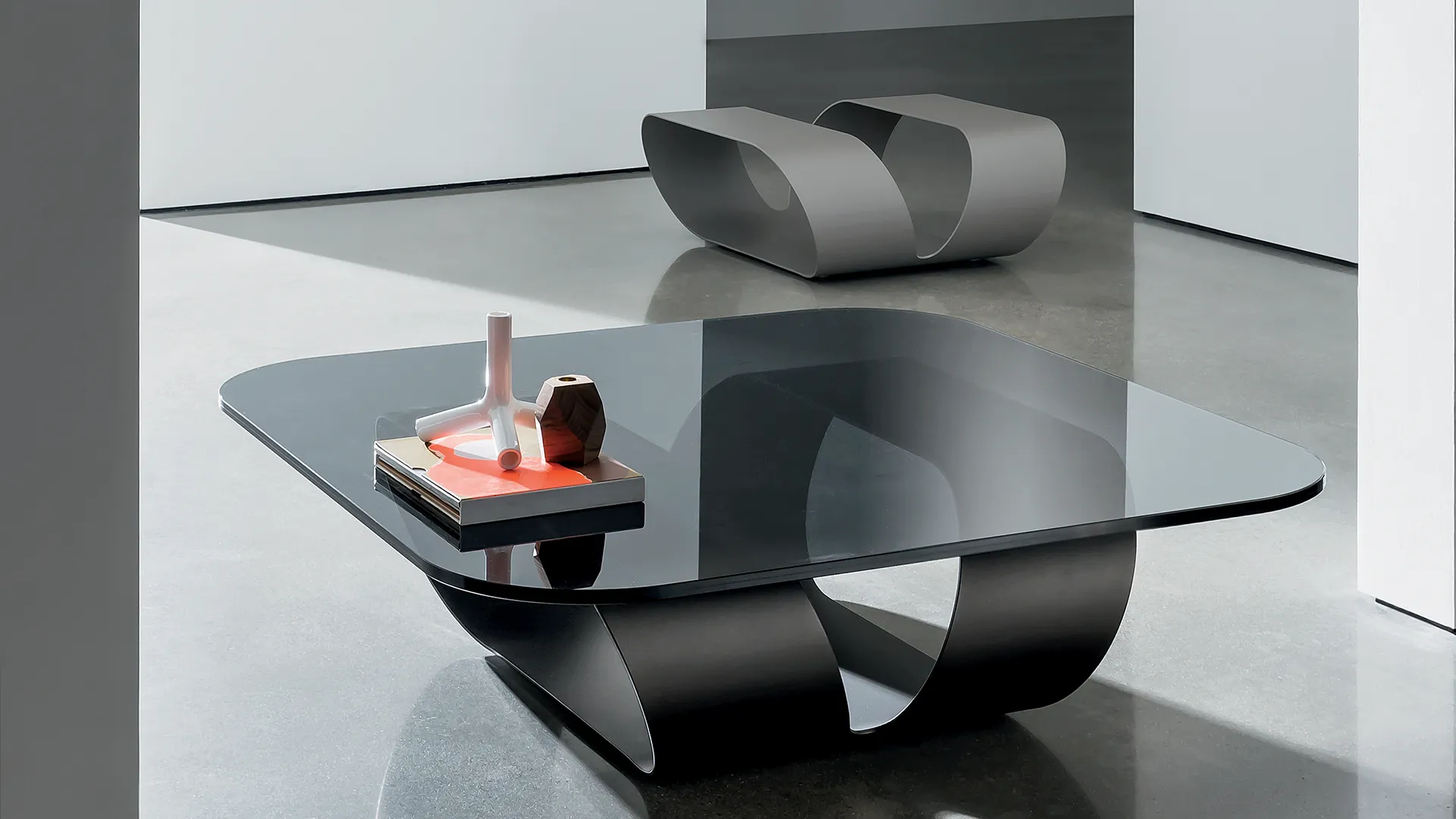 Tavolino in vetro sagomato dalla forma quadrata con piano in vetro fumè e struttura composta da un'unica lastra di metallo curvata di colore grigio 